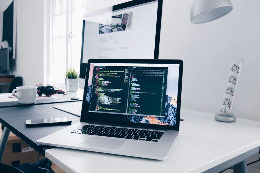 a MacBook Pro displaying lines of colorful code on a dark-themed text editor. Behind it, a larger external monitor shows a webpage. The white desk also holds a smartphone, a white coffee cup, a pair of black headphones, a desk lamp, and a small plant. A window in the background allows natural light to enter the room, which has a clean and organized aesthetic.