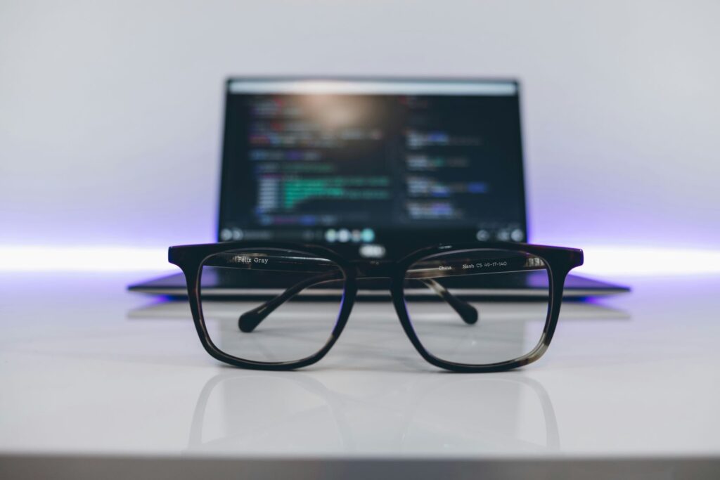 Glasses sit in-focus in front of an out of focus laptop that appears to have a code editor open.