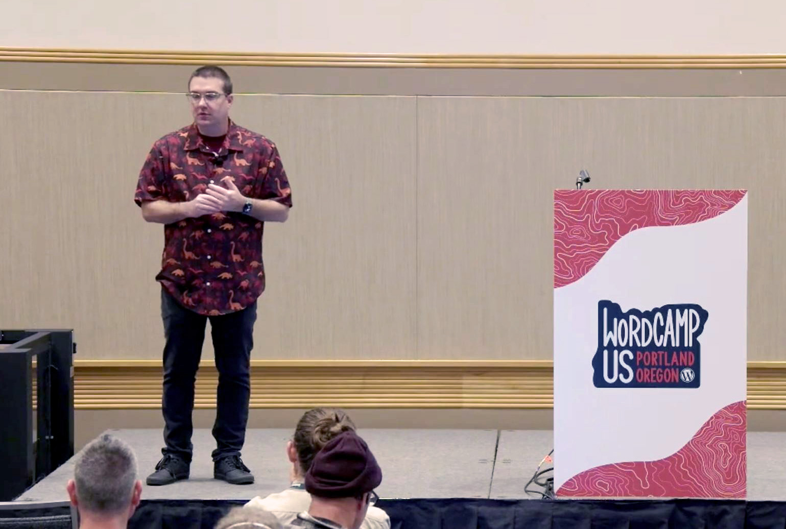 Gary is wearing a red dinosaur-patterned shirt, black jeans, and black sneakers stands on stage at WordCamp US in Portland, Oregon in 2024. He is gesturing with his hands while addressing an audience. A podium with a white and red design featuring the ‘WordCamp US Portland Oregon’ logo is positioned to his left. The audience is visible in the foreground.